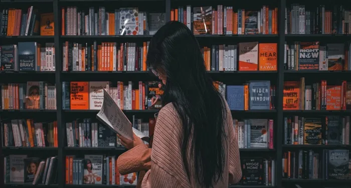 tan-skinned-person-with-long-black-straight-hair-reading-a-book-in-front-of-a-book-stores-shelves.jp_.webp